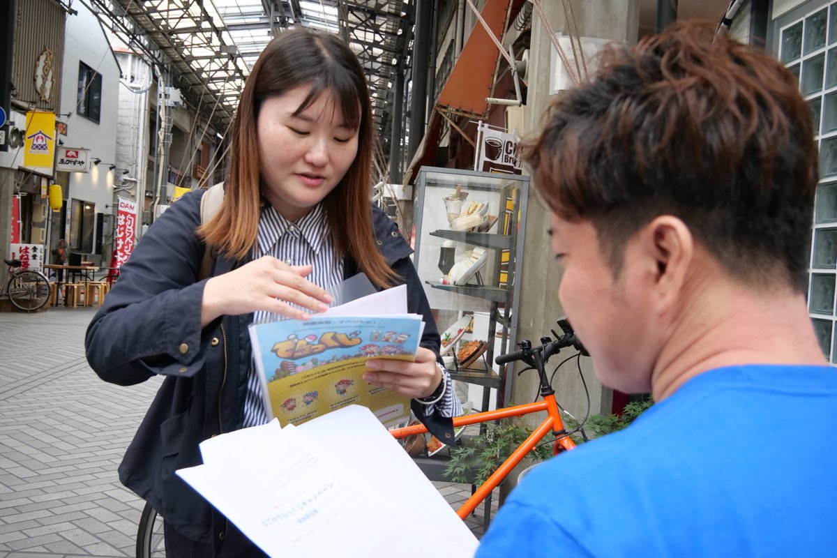 商店街の店先で、店舗の方と一緒にデジタルスタンプラリーのチラシを見る高木さん