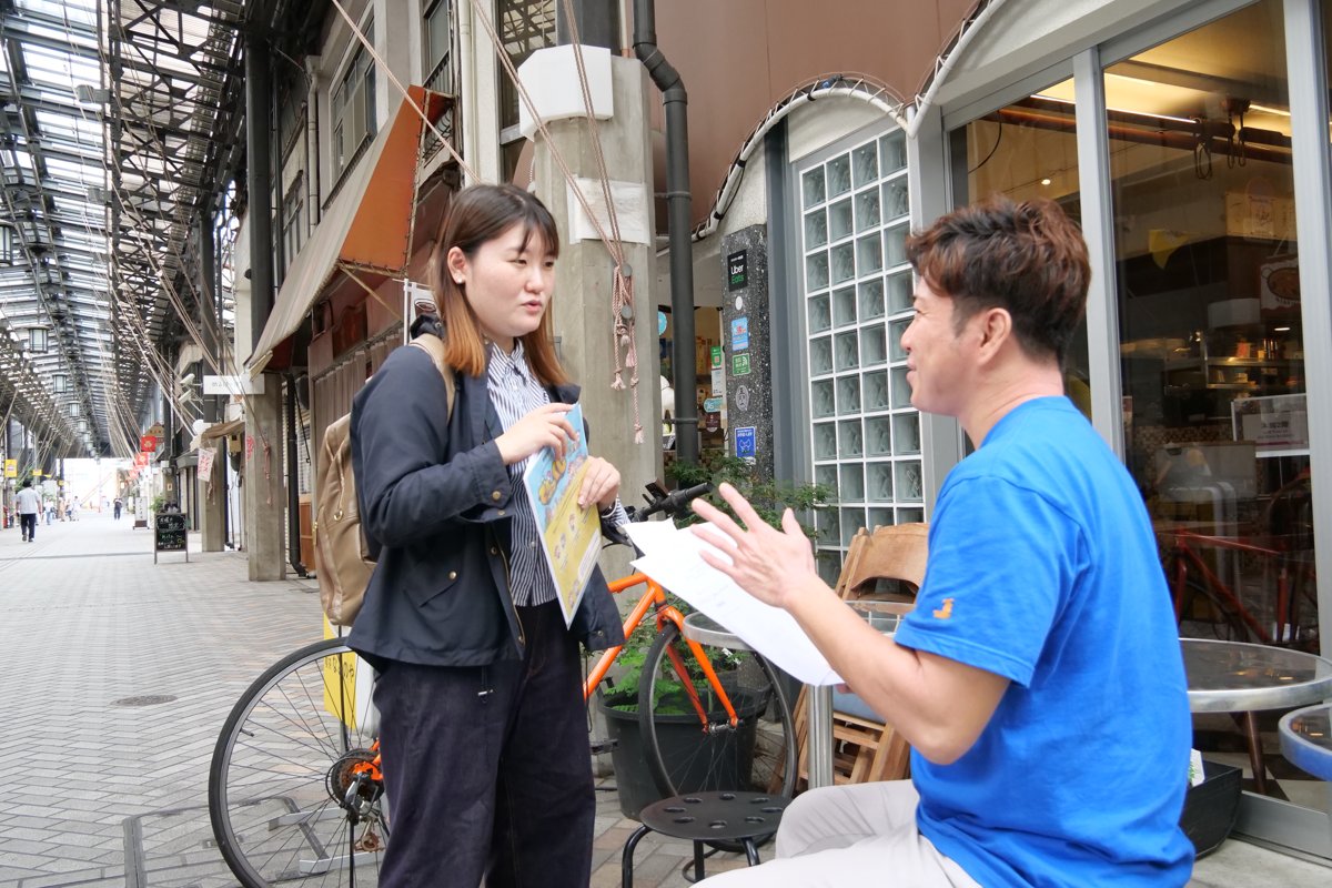 商店街の店先で、店舗の方の話を聞きながらデジタルスタンプラリーのチラシを説明する高木さん